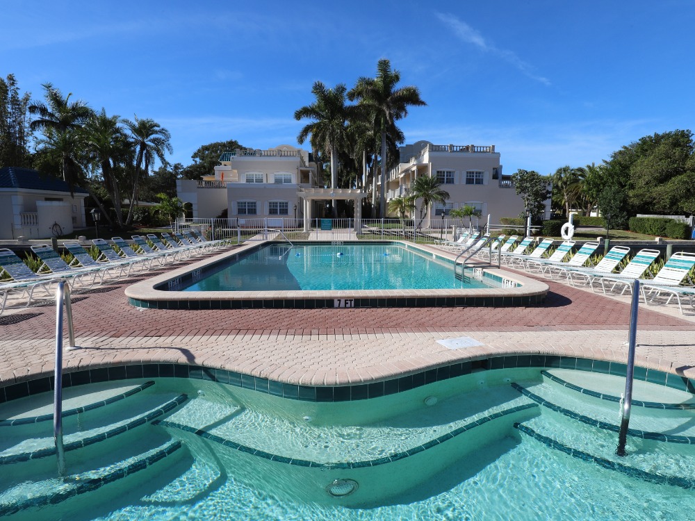 A sunny pool area with a large swimming pool, surrounded by lounge chairs and palm trees, against a clear blue sky.