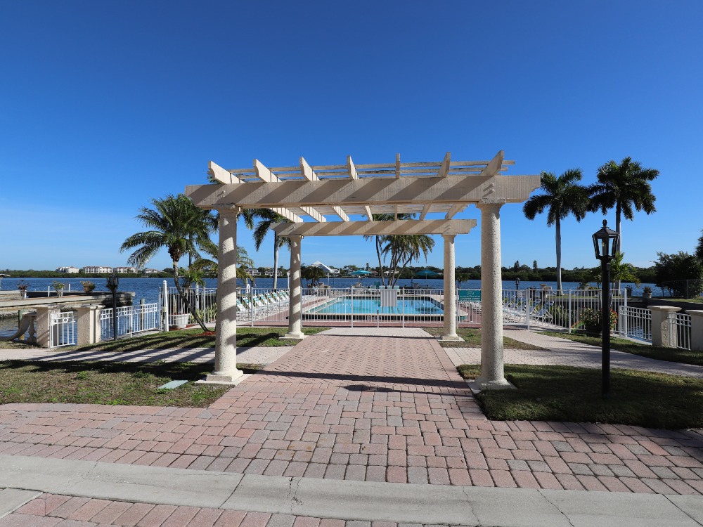 A sunny outdoor area featuring a pergola, palm trees, and a pool by the water, with a clear blue sky above.