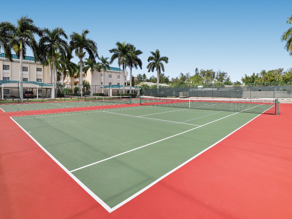 A tennis court with green and red surfaces, surrounded by palm trees and buildings in a clear blue sky.