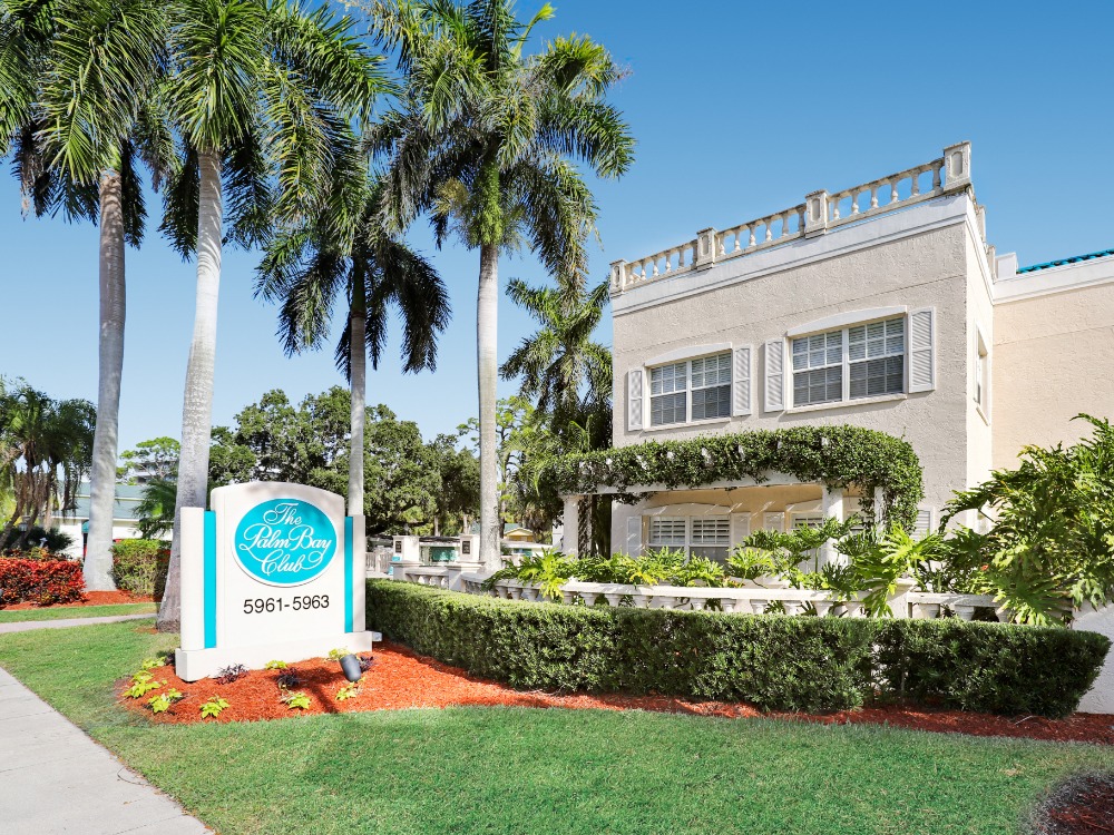 A tropical building with palm trees and lush greenery, featuring a sign for The Palm Bay Club. Bright blue sky above.