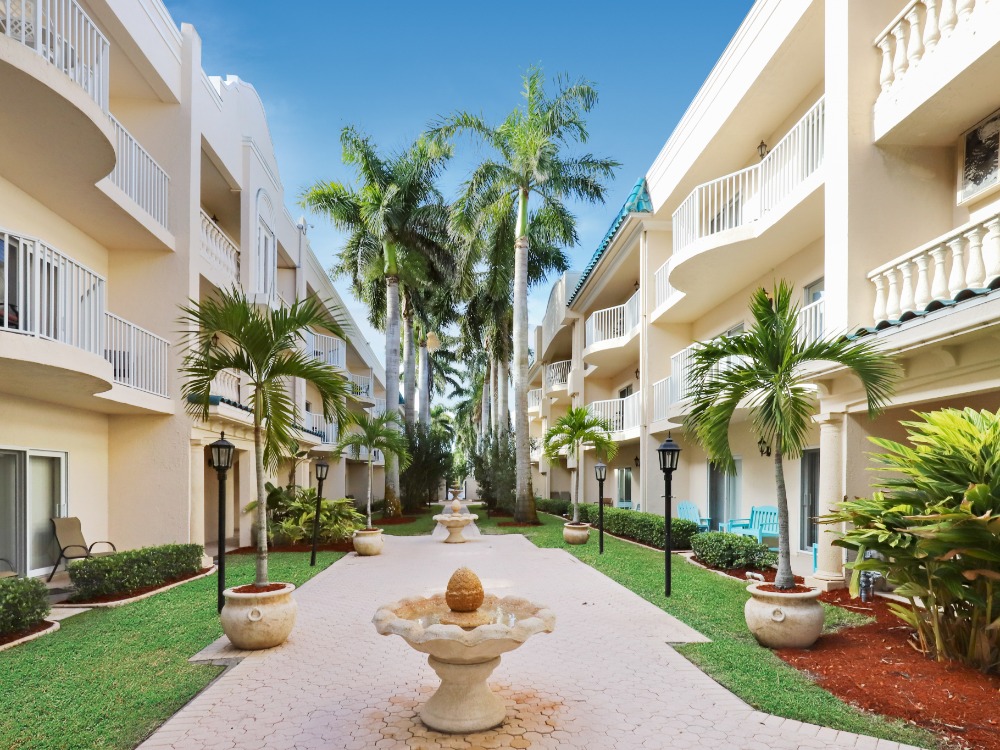 A serene courtyard with palm trees, a fountain, and balconies of a two-story building under a clear blue sky.