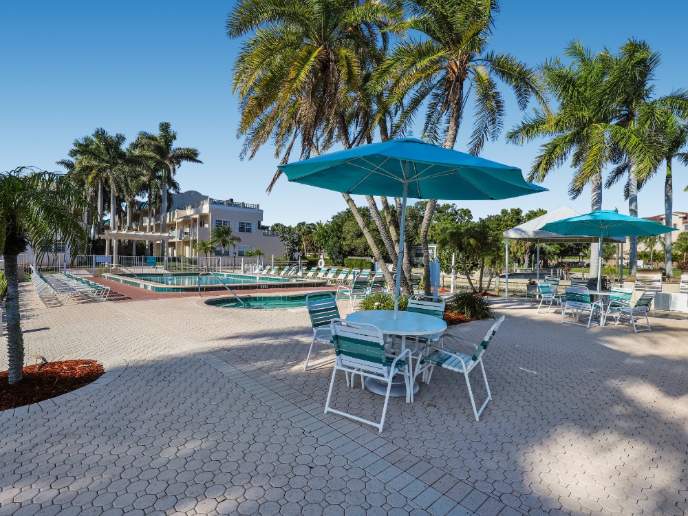 A sunny pool area with palm trees, lounge chairs, and tables under blue umbrellas. A building is visible in the background.