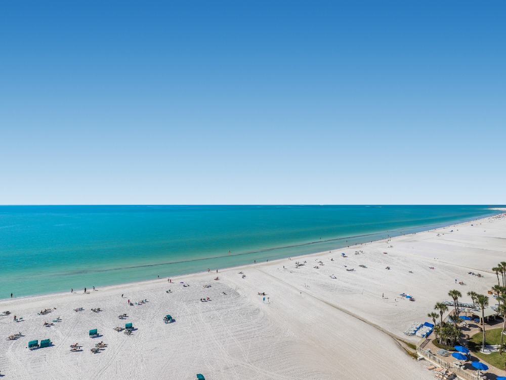A serene beach scene with white sand, turquoise water, and a clear blue sky. Sun loungers are scattered along the shore.