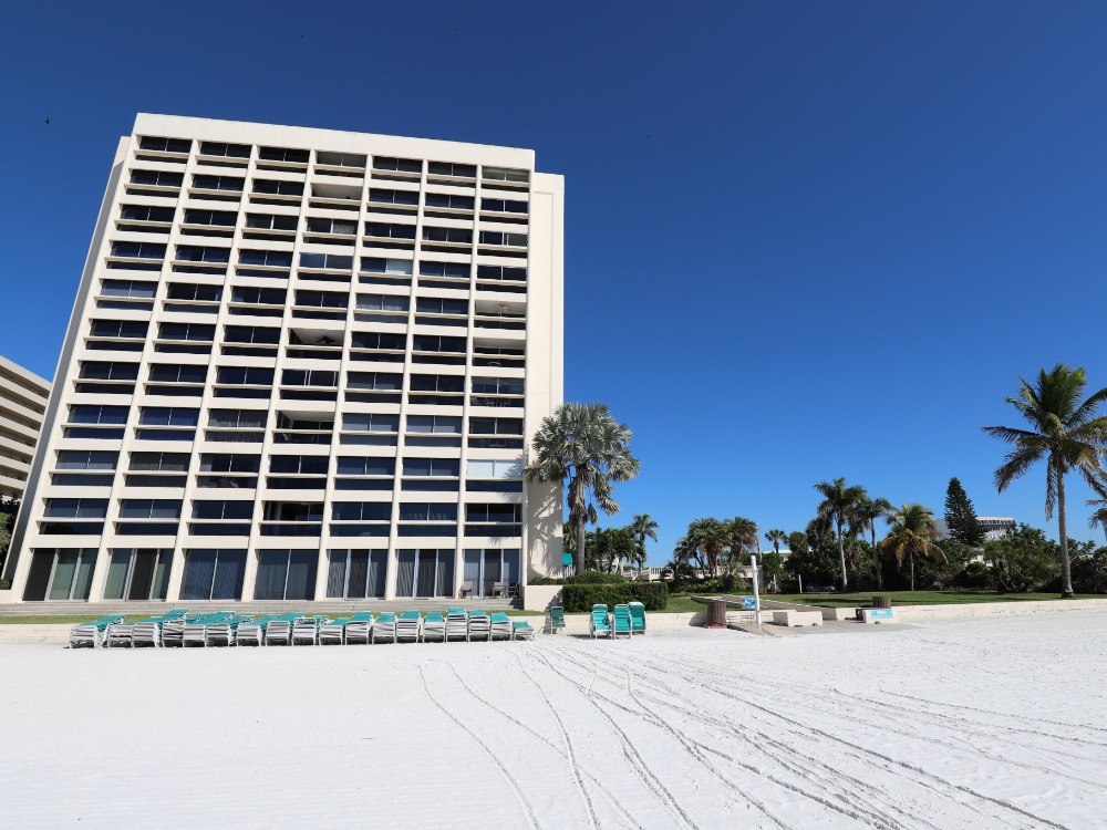A beachfront hotel with large windows, surrounded by palm trees and beach chairs on white sand under a clear blue sky.