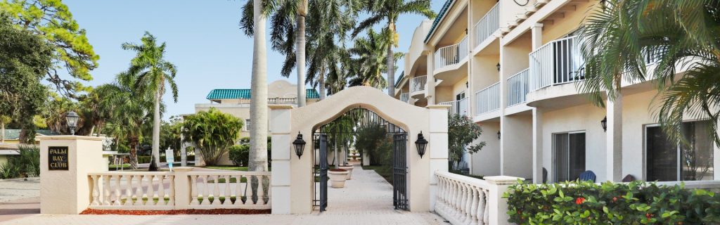 A gated entrance to a tropical resort with palm trees, a pathway, and balconies visible on the buildings.