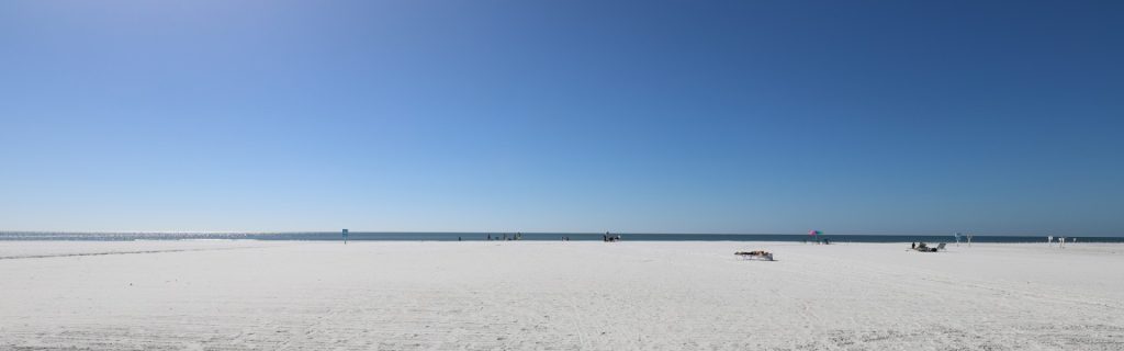A serene beach scene with white sand, a clear blue sky, and gentle waves in the distance. A few people are enjoying the shoreline.