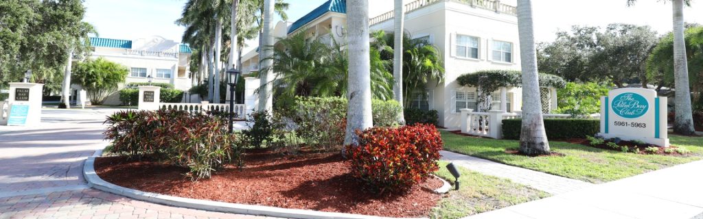 Entrance to the Palm Bay Club featuring palm trees, colorful landscaping, and a welcoming sign.