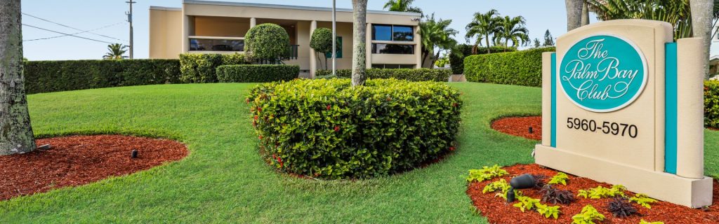 A landscaped entrance featuring a sign for "The Palm Bay Club," surrounded by green grass and decorative shrubs.
