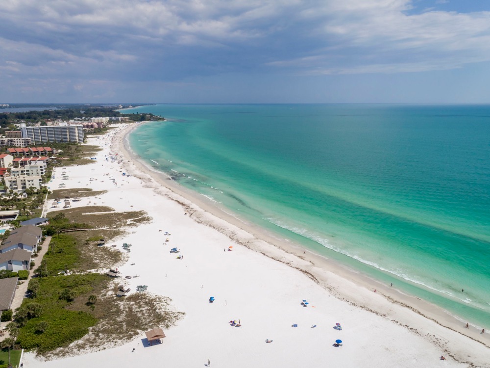 Aerial view of a sandy beach with turquoise waters, dotted with umbrellas and people enjoying the sun. Coastal buildings in the background.