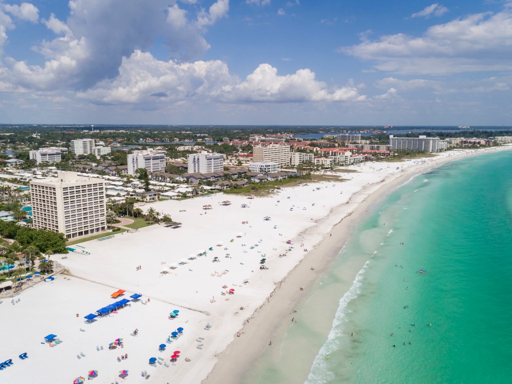Aerial view of a beach with white sand, turquoise water, and scattered umbrellas, alongside a coastal town with buildings.