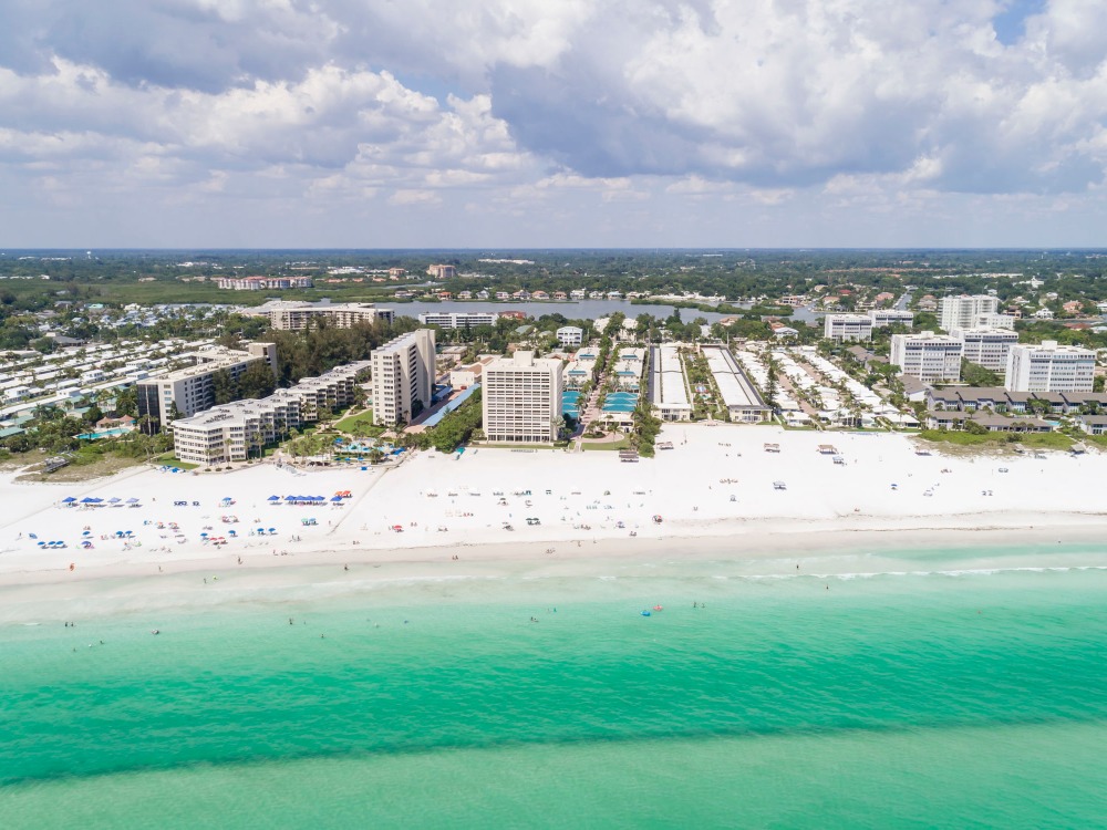 Aerial view of a beach with white sand, turquoise water, and several buildings along the shore. Sunbathers and umbrellas are visible.