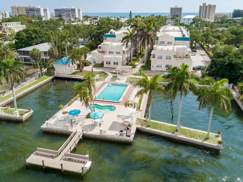 Aerial view of a waterfront property featuring a pool, palm trees, and nearby buildings with a glimpse of the ocean in the background.