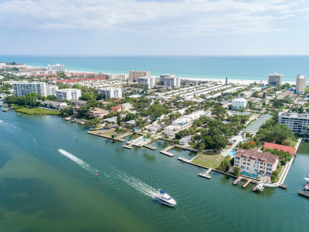 Aerial view of a coastal area with residential buildings, waterways, and a beach in the background. A boat is navigating the water.