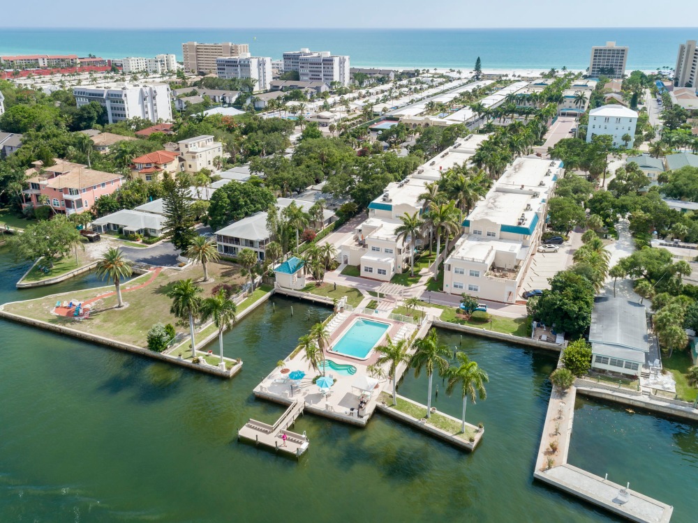 Aerial view of a coastal area featuring residential buildings, palm trees, a pool, and waterways leading to the ocean.