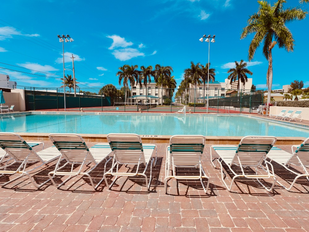 A sunny pool area with lounge chairs, palm trees, and a clear blue sky. Tennis courts are visible in the background.