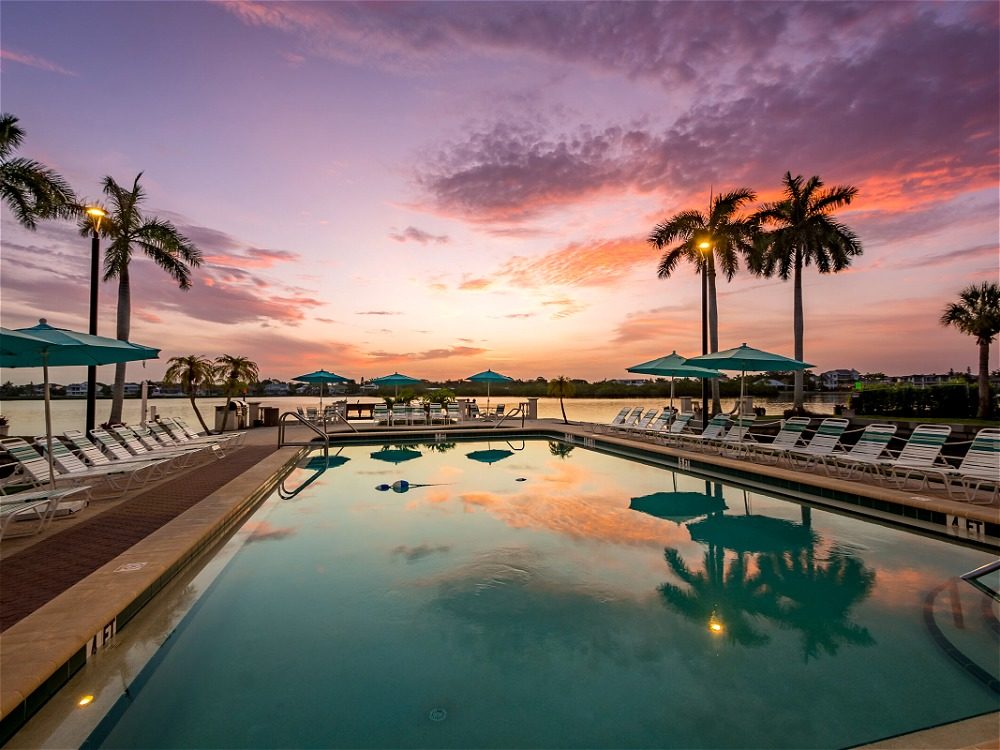A serene poolside scene at sunset, featuring palm trees, lounge chairs, and umbrellas, reflecting vibrant colors in the water.