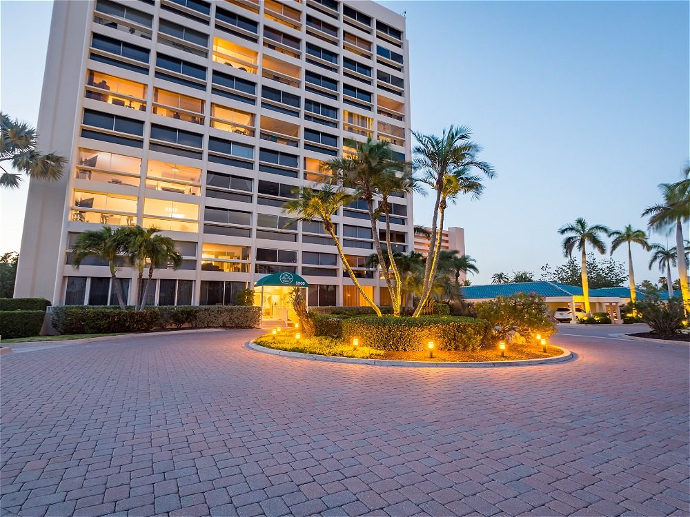 A modern building with illuminated windows, surrounded by palm trees and a circular driveway at dusk.