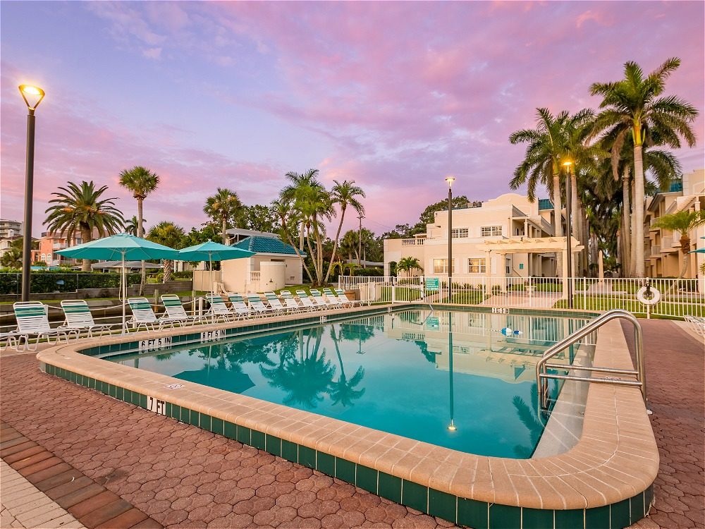A serene pool area surrounded by palm trees, with lounge chairs and umbrellas, under a colorful sunset sky.
