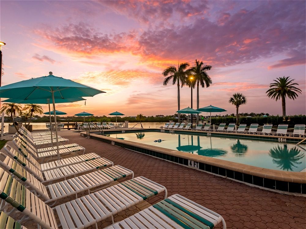 A serene poolside scene at sunset, featuring lounge chairs, umbrellas, and palm trees against a colorful sky.