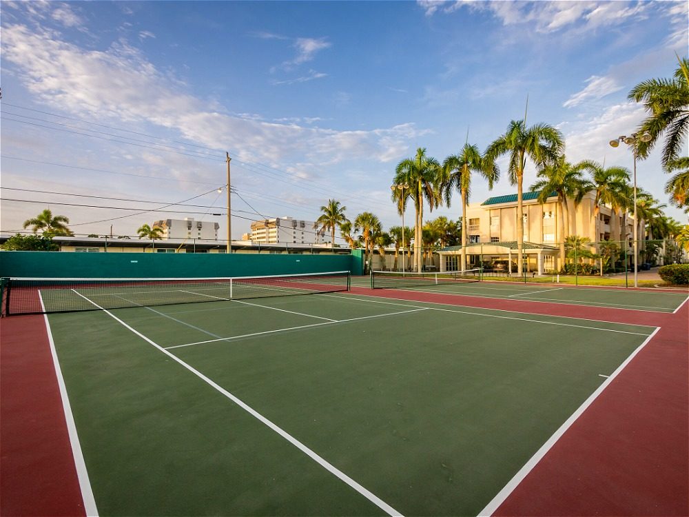 A tennis court with green and red surfaces, surrounded by palm trees and buildings under a clear blue sky.