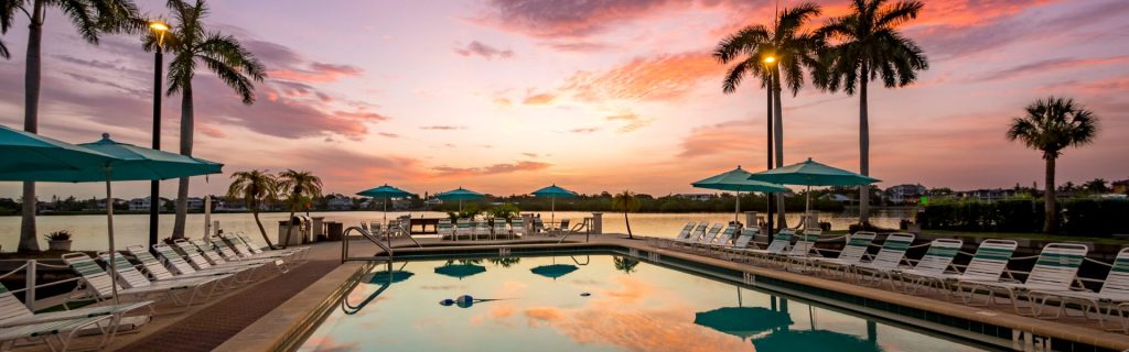 A serene poolside scene at sunset, featuring lounge chairs, umbrellas, and palm trees, reflecting on the water.