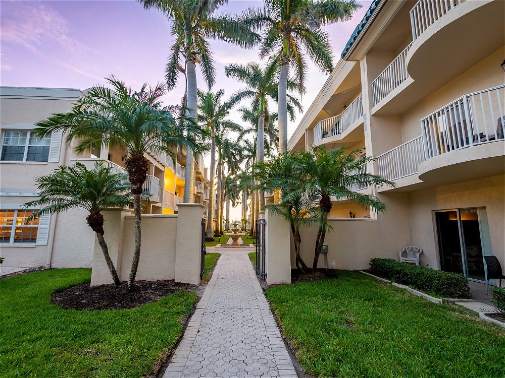 A pathway lined with palm trees leads through a tropical courtyard, flanked by two buildings with balconies.