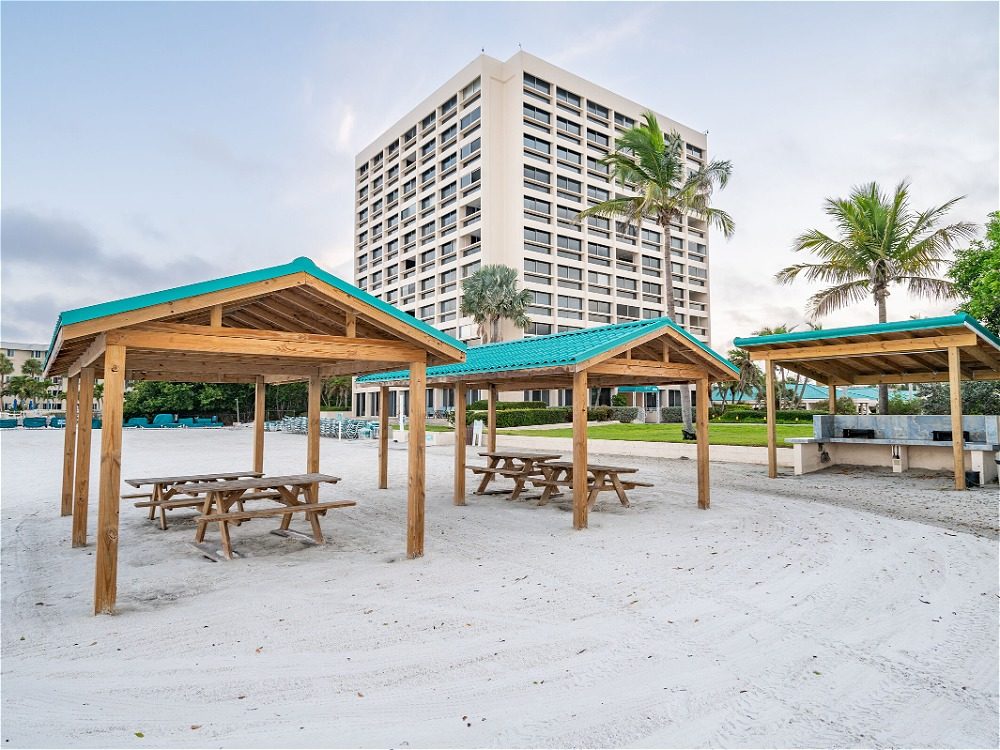 Beachside pavilions with picnic tables in front of a tall hotel, surrounded by palm trees and sandy ground.