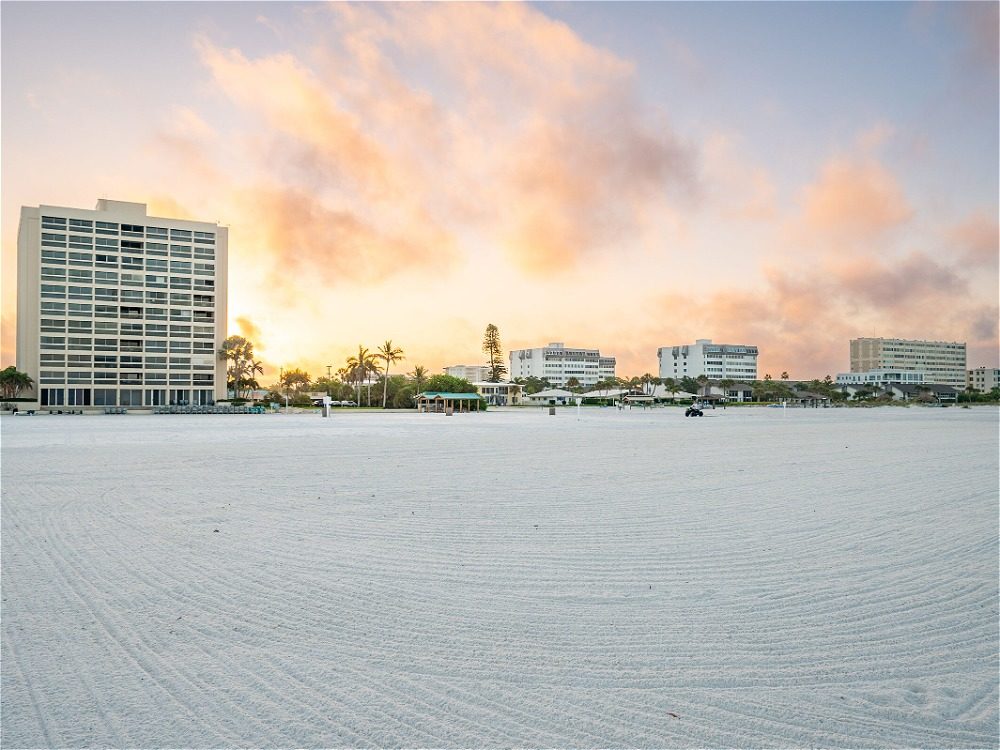 A beach scene at sunset with several buildings in the background and smooth, sandy foreground.