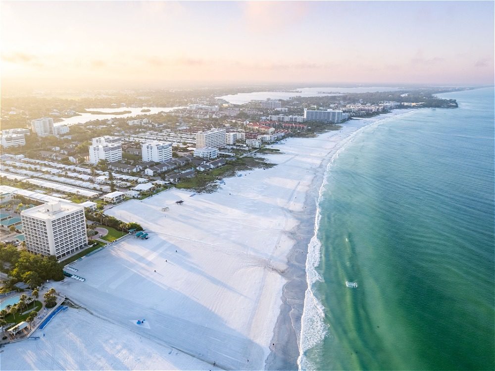 Aerial view of a beach with white sand and turquoise water, lined with buildings and greenery under a soft sunset sky.