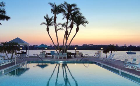 jqg_1556313468-1 A serene poolside scene at sunset, featuring palm trees, lounge chairs, and a calm water view.
