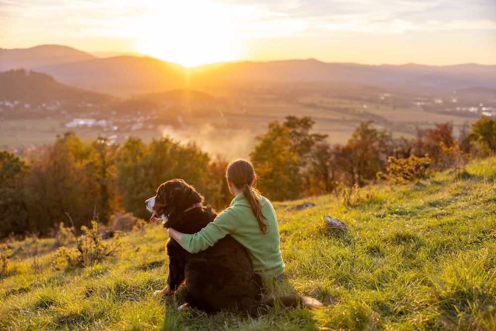 woman and dog sit on a mountain watching sunset over peaks